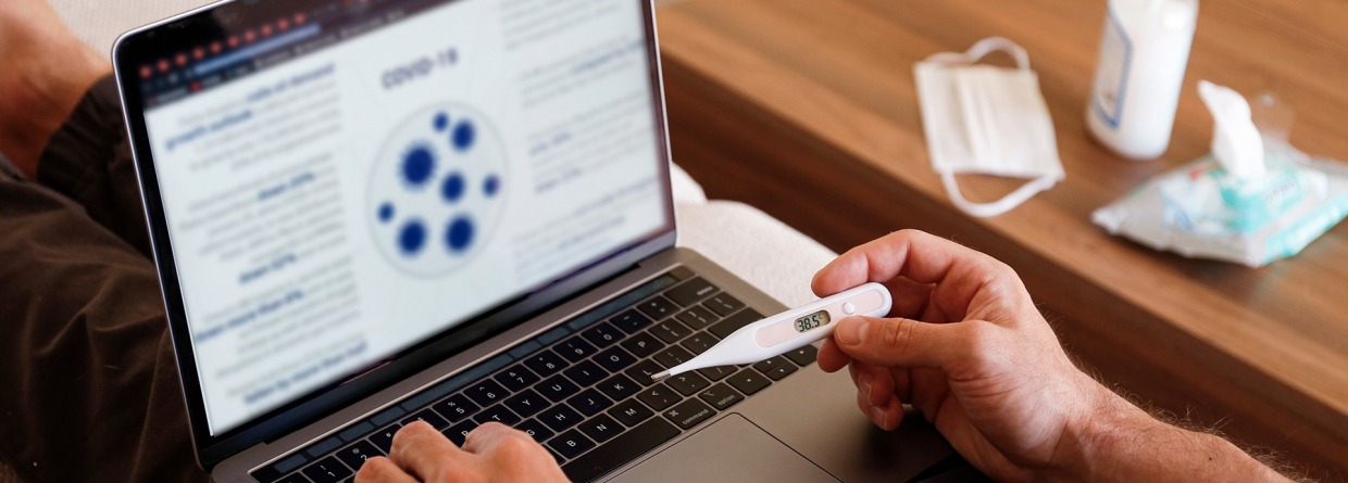 Beeld van ziek mens die vanuit huis werkt. Laptop op schoot met thermometer in hand. Op de tafel ligt een mondkapje, tissues en desinfectie handgel